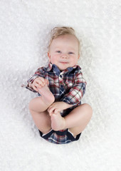 a six-month-old blond boy with blue eyes in a checkered shirt bodik lies on his back playing with his legs, smiling cute, on a white background