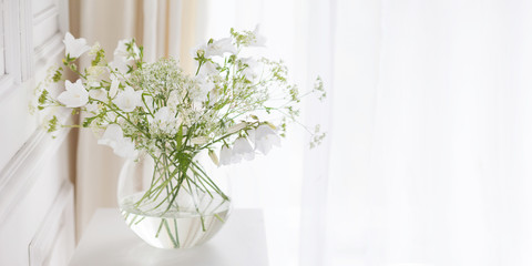 Bouquet of gentle bells in vase. Morning light in the room. Soft home decor, glass vase with white flowers on  white wall background and on wooden table. Interior. Greeting card. Copy space. Banner