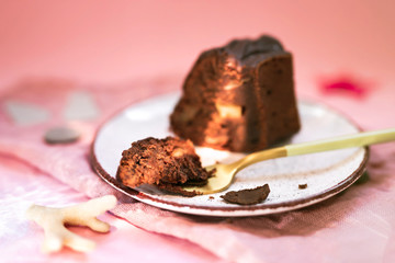 A slice of a chocolate bundt cake with apples on a white plate, a piece of cake on a gold fork near. Pink napkin and background. Patches of light on the scene.