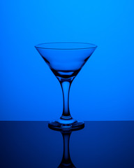 Glass goblet without wine on a thin leg stands on a mirror surface. Back light. Dark blue background. Vertical orientation.