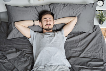 Young bearded man lying with hands behind head while sleeping alone in bed