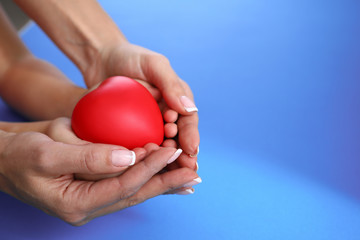 Close-up of mother and child holding red heart in hands on blue backdrop. Copy space in right side. Motherhood and protection. Family relationship charity and adoption concept