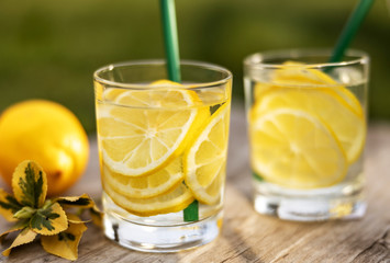 Summer drink, glasses of water filled with slices of lemons, stand on a wooden board on the lawn in sunny warm day, the background is blurred, shallow depth of field, selective focus.