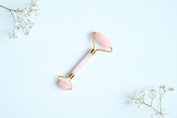 Rose quartz facial massage roller on blue background with gypsophila flowers. Facial massager with...