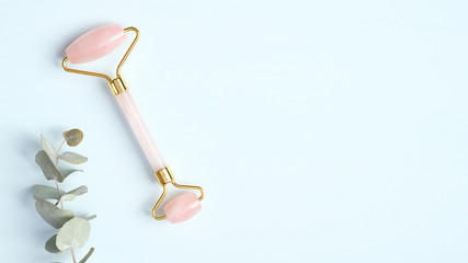 Rose quartz jade roller and eucalyptus leaf on blue background. Facial massage roller, anti-aging, anti-wrinkle beauty skincare tool