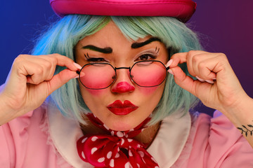 Close-up portrait of a clown girl with blue hair in a pink hat, pink dress and glasses. A clown...