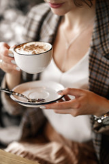 Close-up of a white cup of coffee and a white saucer in the hands of a beautiful girl of European race. She's wearing a checkered jacket.