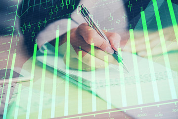 A woman hands writing information about stock market in notepad. Forex chart holograms in front. Concept of research. Multi exposure