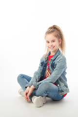 Young girl wearing denim jacket, red shirt, jeans and white sneakers sitting cross-legged on floor