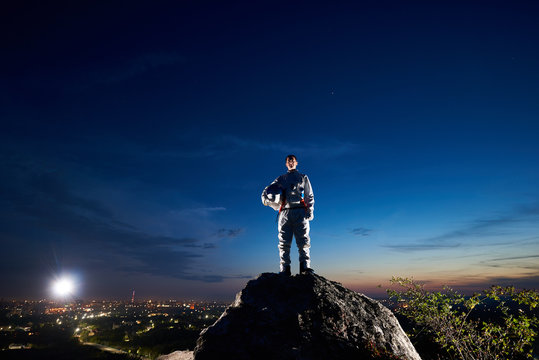 Brave astronaut standing on top of rocky hill with beautiful blue sky, city lights on background. Handsome spaceman in white space suit holding helmet and looking at camera. Concept space travel.