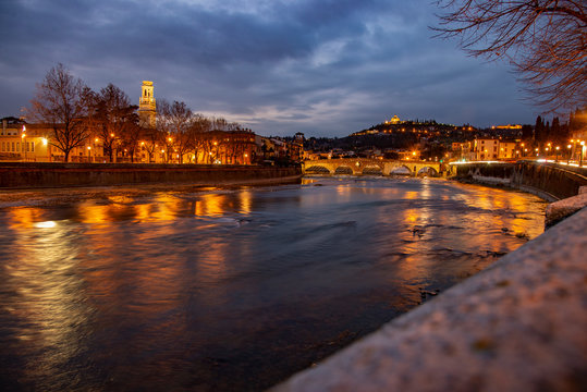 Night photo along the Adige river, Verona, Italy. Stone bridge with the bell tower of the cathedral and the sanctuary of the madonna of Lourdes.