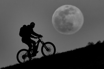 Fototapeta premium Mountainbiker rides his Bike in front of the full moon (super moon) during moon rise and sunset.
