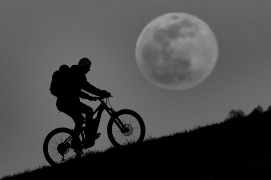 Mountainbiker rides his Bike in front of the full moon (super moon) during moon rise and sunset.
