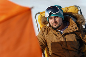 The guy sits in the winter on a yellow cot under an umbrella and looks at the camera