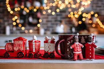 On the table are beautiful mugs, as well as kitchen utensils in the form of a train. The kitchen is decorated for Christmas. Next to the mugs is a ginger cookie in the form of men in a Santa costume.