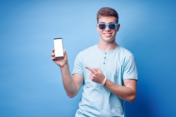 Copy space for app. Handsome young man pointing on screen of smartphone. Studio portrait on blue background.