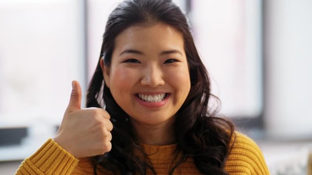 people, ethnicity and race concept - portrait of happy smiling asian young woman showing thumbs up at home