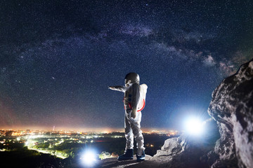 Obraz premium Male spaceman pointing finger at night city in valley while standing on rocky mountain under fantastic night sky with stars and Milky way. Concept of cosmonautics and space traveling.