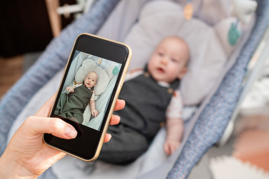 Mother takes a picture with her son by phone. The woman will send photos to grandparents via email. Mom is spending time with her cute baby boy at home office. Mother's love. Maternity leave content.
