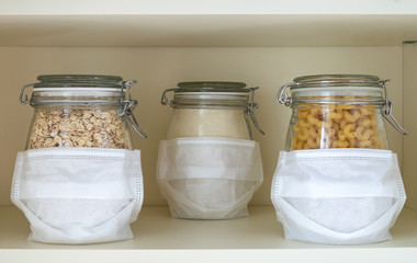 Glass jars with stocks of products in masks stand at a distance from each other on a shelf in a cupboard. Social distancing, wearing a mask, COVID-19 concept. Horizontal orientation, selective focus