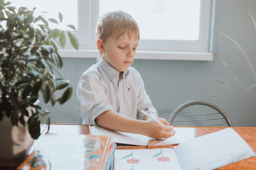 The boy writes in a notebook at home during CoVid-19 quarantine, distance learning online with a laptop, a child doing homework for school. Children stayed at home.