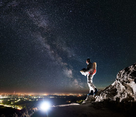 Fototapeta na wymiar Cosmonaut standing on rocky hill under beautiful night sky with stars and Milky way. Space traveler in space suit holding helmet and looking at night city lights. Concept of cosmonautics and nighttime