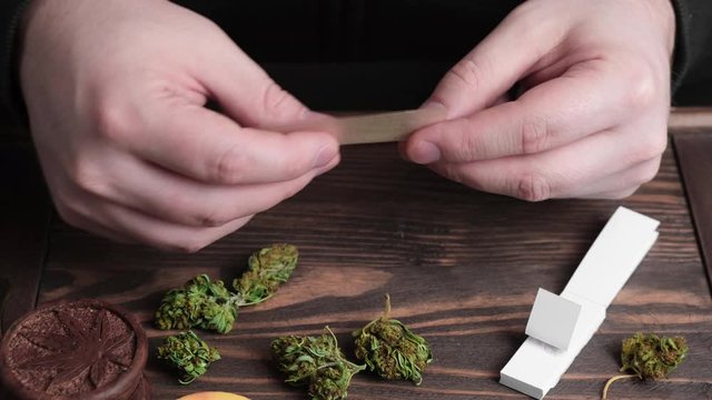 Man prepair cigarette paper for hand made cannabis joint over the wooden desk with marijauna buds and paper filters on it.