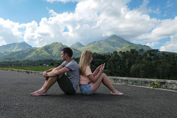 Naklejka na ściany i meble Girl and boy sitting on the road to the mountains. Green mountains with blue sky and clouds. Couple in the nature.