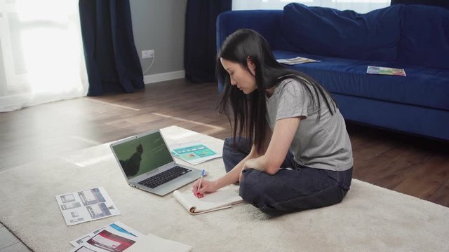 Side view of female Asian student sitting on floor at home, studying online watching video conference with teacher and taking notes in notepad
