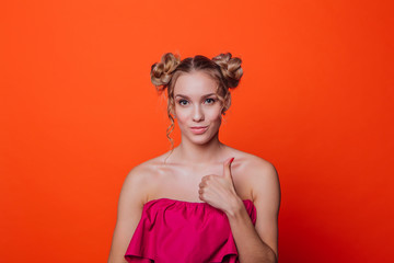 The girl in a red dress on a orange background in the studio. Blonde girl with two hair knots showing thumb up, looking to the camera and smiling.
