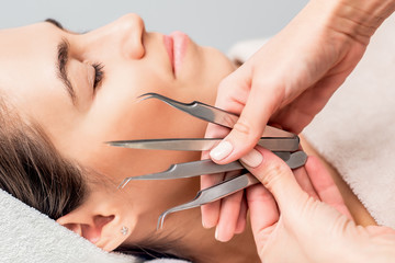 Obraz na płótnie Canvas Hands of cosmetologist holds tweezers for eyelash extensions on background of face of young woman, close up.
