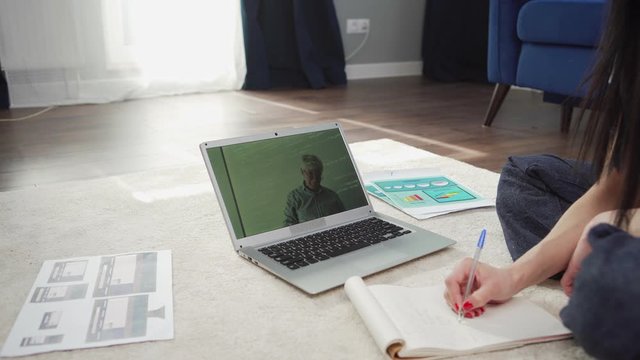 Tilt up side view of female Asian college student sitting on floor at home, studying online watching video conference with teacher and taking notes in notepad