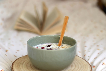Breakfast bowl with milk, oats, chia seeds and berries, served on a bed with floral sheets. Open book in the background. Selective focus.