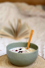 Breakfast bowl with milk, oats, chia seeds and berries, served on a bed with floral sheets. Open book in the background. Selective focus.