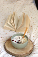 Breakfast bowl with milk, oats, chia seeds and berries, served on a bed with floral sheets. Open book in the background. Selective focus.