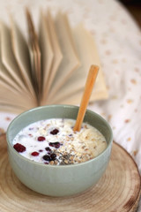 Breakfast bowl with milk, oats, chia seeds and berries, served on a bed with floral sheets. Open book in the background. Selective focus.
