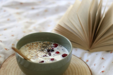 Breakfast bowl with milk, oats, chia seeds and berries, served on a bed with floral sheets. Open book in the background. Selective focus.