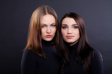Two young girlfriends are standing together. Both are in black clothes and are looking at the camera. Black background