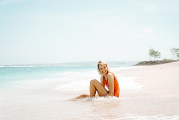 Fototapeta na wymiar Blonde woman sitting on a white sandy beach wearing the red swimsuit. She is smiling at the camera and enjoying hot summertime.