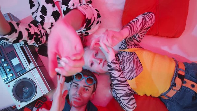 Top view shot of three young dancers in stylish outfits lying on the floor with retro cassette player, posing for camera and performing vogue hands movements under pink lighting