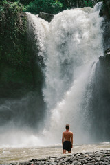 A young man standing under a big / tall waterfall in Indonesia.