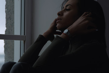 Depressed sad young african american woman feels anxiety stress sit alone by the window at home....