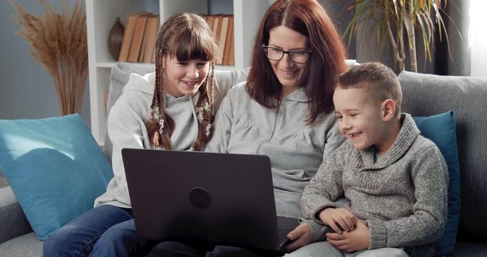Smiling woman in eyeglasses with two children relaxing on comfortable couch and using laptop. Mother with her son and daughter enjoying free time with computer at home.