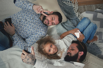 Top View Of Male Couple And Their Five Year Old Daughter Having Fun Taking Selfies With Smart Phone Camera App While Making Faces. Same Sex Couple With Daughter Having Fun