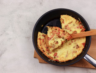 Frittata with potatoes, green peas, cheese and sausagesin in a frying  pan on a wooden cutting board on a on a light gray background. Top view, flat lay