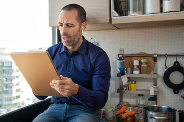 Uomo moro con camicia blu lavora in modalità smart working in cucina  con il suo  tablet