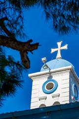 Beautiful white cross of Orthodox church of Monastery of St. Nicholas, lake Vistonida, Porto Lagos, Xanthi region, Northern Greece, bell tower partial view against clear blue sky, framed by trees