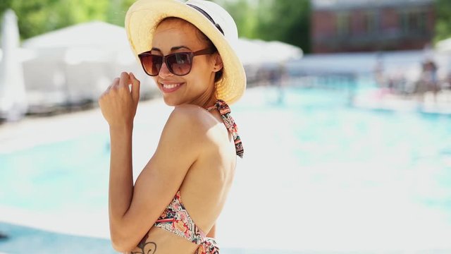 slow motion Beautiful woman in bikini, hat and sunglasses joyfully smiling and posing on poolside background. poolside leisure and relaxation at a luxury spa hotel