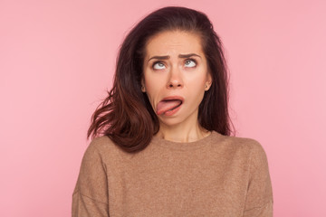 Fototapeta premium Portrait of funny silly young woman with brunette hair looking cross eyed and sticking tongue out, making dumb brainless face, disobedient goofy expression. studio shot isolated on pink background