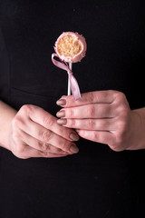 Cake pops in women's hands on a black background. Dessert in pink chocolate cream with powder and bow.